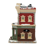 Porcelain lighted building for a Lemax tabletop display. Designed to look like a red, gray and brown brick building. There's snow on the roof and the sidewalk. There are signs advertising croissants, coffee, espresso and sandwiches. Inside the window you can see a man sitting at a table. There's a dog sitting under the window on the sidewalk.
