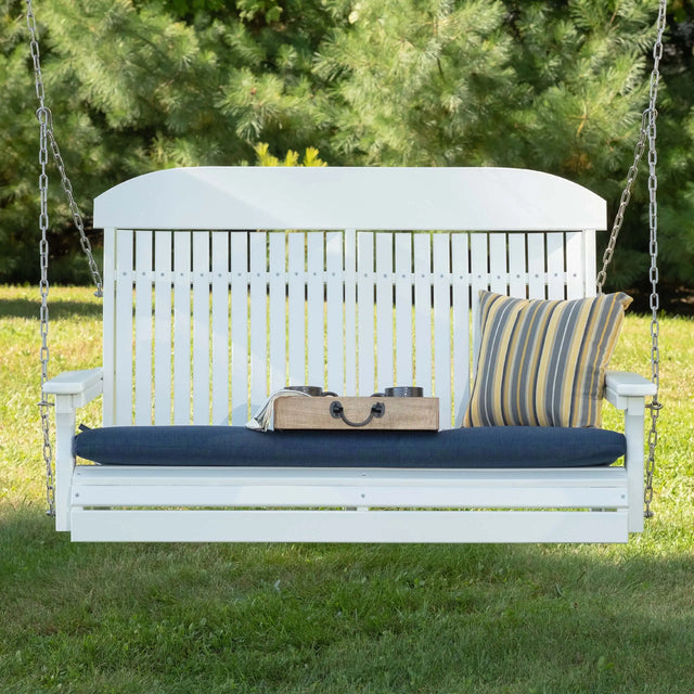 This is a lifestyle image showing the White swing in a grassy backyard. There's a blue seat cushion and a yellow and blue striped throw pillow.