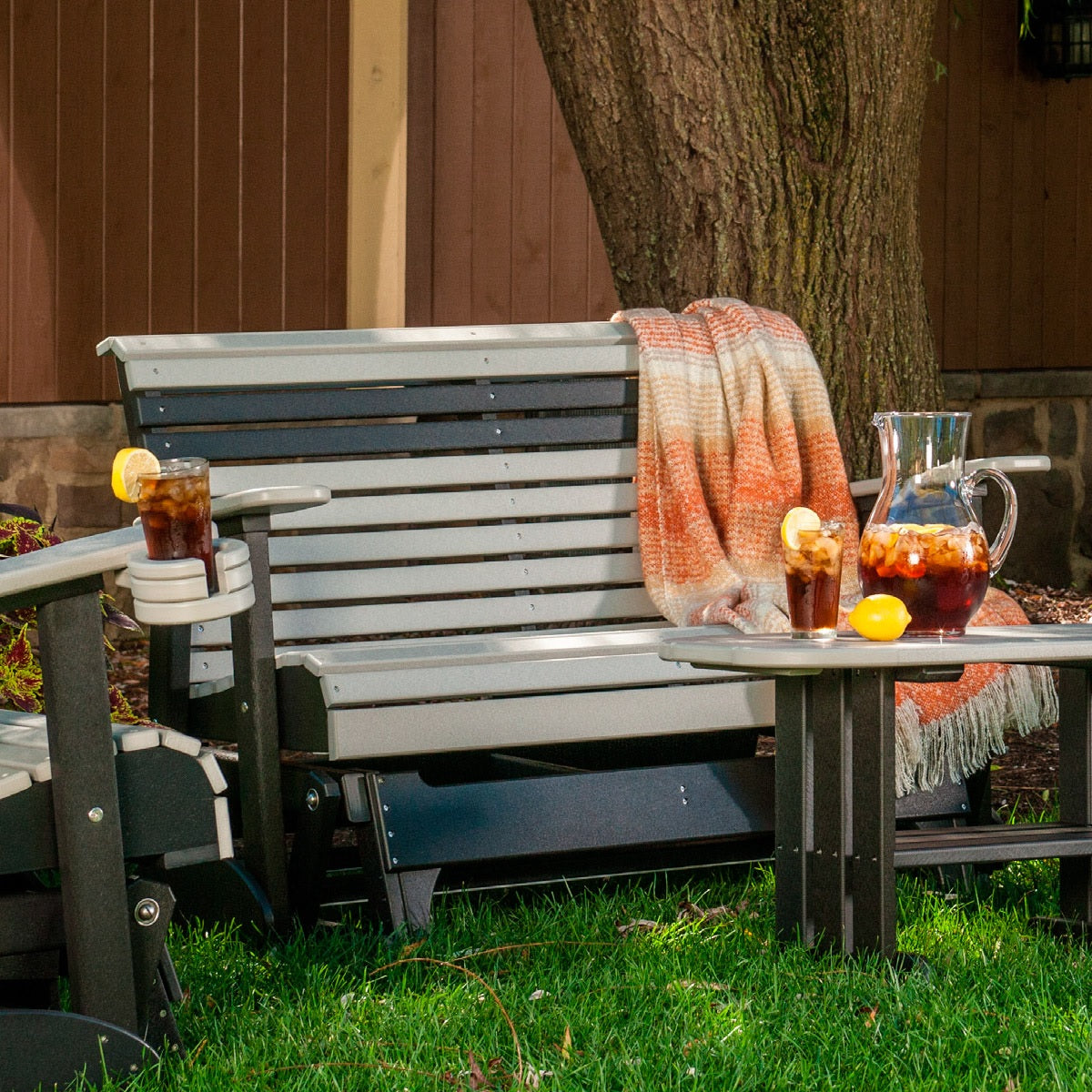 This is a lifestyle image of the Dove Gray and Black colored glider bench in a grassy yard. There's an orange and white colored blanket draped over the side.