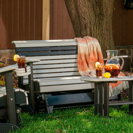 This is a lifestyle image of the Dove Gray and Black colored glider bench in a grassy yard. There's an orange and white colored blanket draped over the side.