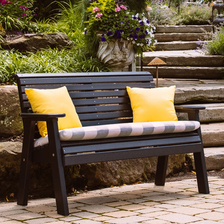 This is the Black colored bench on a stone patio with two yellow throw pillows and a seat cushion on it.