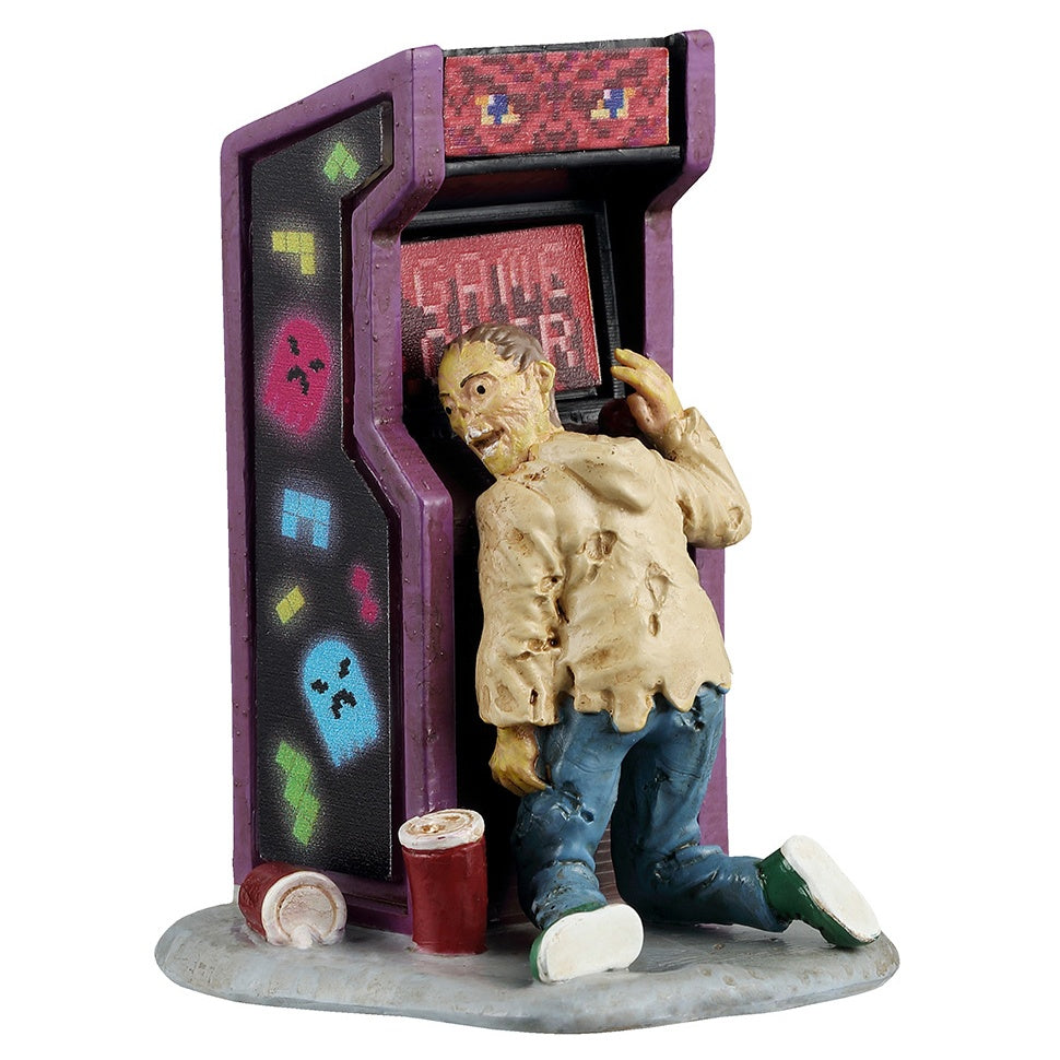 This image shows what this Lemax Spooky Town figurine looks like on a white background. It was designed to look like a zombie leaning up against a reto-looking arcade game. The screen reads "GAME OVER." There are two drinks discarded on the ground. The sides of the game are decorated with neon colored Tetris pieces and neon colored Pac-Man style ghosts.
