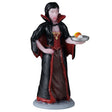 This image shows what this Lemax Spooky Town figurine looks like on a white background. It's designed to look like a woman-vampire holding a silver plate with candy corn on it. She has black hair and is wearing a black and red robe over a red and brown dress. She's wearing a gold necklace and has blood coming off of her fangs.