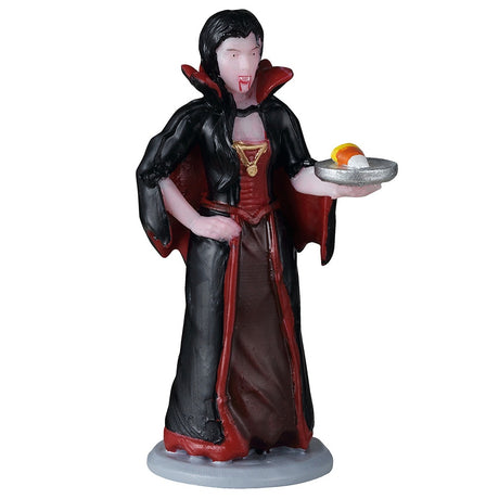 This image shows what this Lemax Spooky Town figurine looks like on a white background. It's designed to look like a woman-vampire holding a silver plate with candy corn on it. She has black hair and is wearing a black and red robe over a red and brown dress. She's wearing a gold necklace and has blood coming off of her fangs.