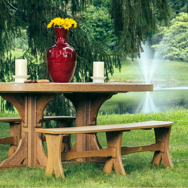 This is a lifestyle image showing the Antique Mahogany colored Table Bench around a matching table in a grassy yard with a small pond with a fountain in the background.