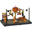 This image shows what this Lemax Spooky Town table accent looks like on a white background. It's designed to look like a space where witches learn how to fly. The instructor and her black cat are taking notes watching two young witches fly in circles. They're flying around a sign that reads "Zoomie Brooms Flying School." There are orange construction cones and brooms all over.