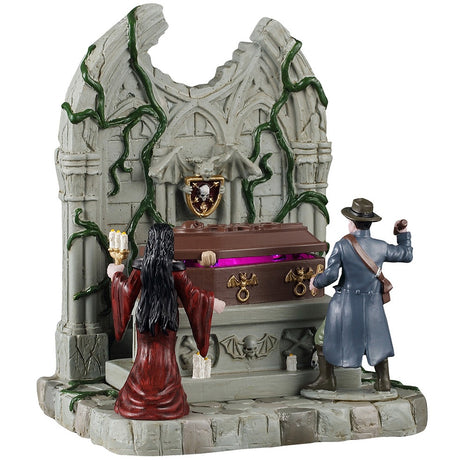 This image shows what this Spooky Town table accent looks like on a white background. This piece was designed to look part of an old-timey mausoleum with green vines growing all up the wall. There's a brown coffin in the center. There's a woman with long black hair wearing a red dress and holding a candelabra. There's a man wearing a long blue coat and a brown hat. He appears to be holding a stake. 