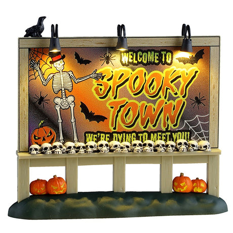This image shows what this Lemax Spooky Town accessory looks like on a white background. It was designed to look like a big, lighted sign. It reads "Welcome to Spooky Town. We're dying to meet you." The sign is decorated with a skeleton wearing a top hat, bats, spiders, spiderwebs and a pumpkin. There's a crow resting on top of the sign and a line of skulls on the bottom of the sign. There are jack-o-lanterns on the ground underneath the sign.
