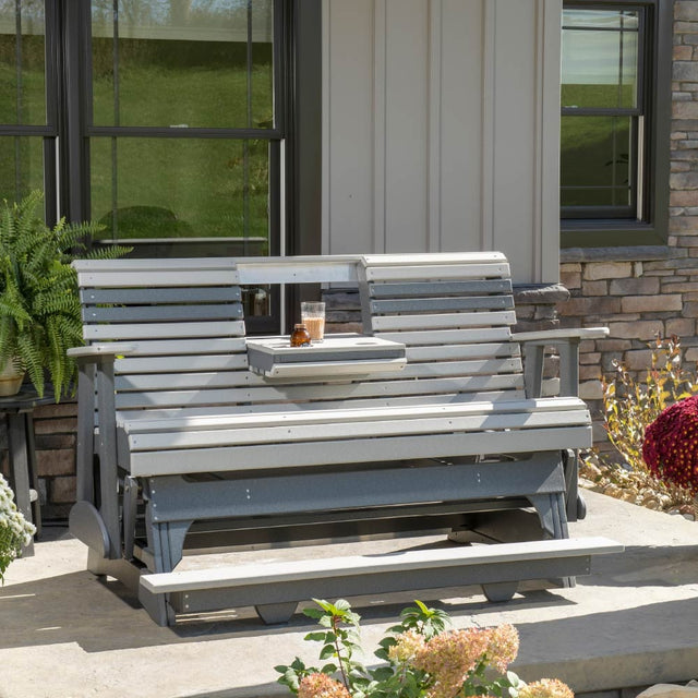 This is a lifestyle image showing the Dove Gray and Slate colored glider bench on a patio in front of a home. The center console is down and there's a class of juice in one of the cupholders.