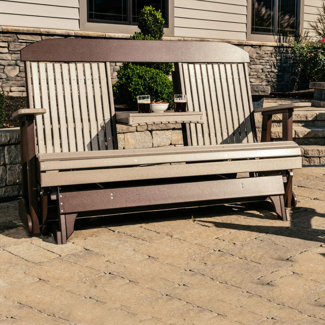 This is a lifestyle image showing the Weatherwood and Chestnut Brown colored classic glider bench on a stone patio in front of a house.
