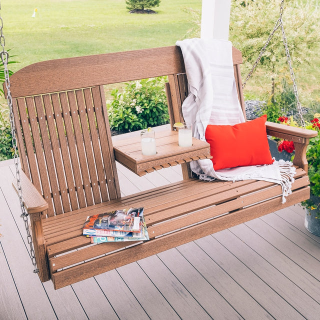 This is a lifestyle image showing the Antique Mahogany porch swing on a wooden porch. There are magazines on one side and a blanket and a red throw pillow on the other side.