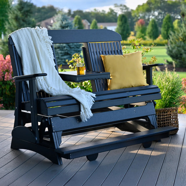 This is a lifestyle image showing the Black colored balcony glider bench on a wooden porch. There's a white blanket draped over the side and a yellow throw pillow on the other side.