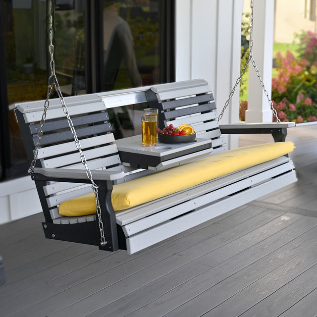 This is a lifestyle image showing the Dove Gray and Black colored porch swing in front of a house. There's a yellow seat cushion on it. The center console is down and there's a glass of juice in the cupholder and a bowl of fruit.