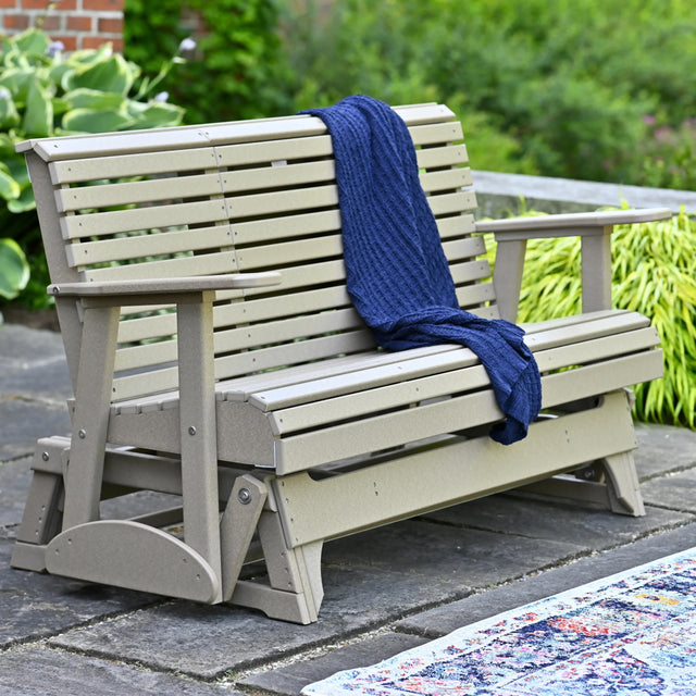 This is a lifestyle image showing the Weatherwood colored glider bench on a stone patio. There's a blue blanket draped over the side.