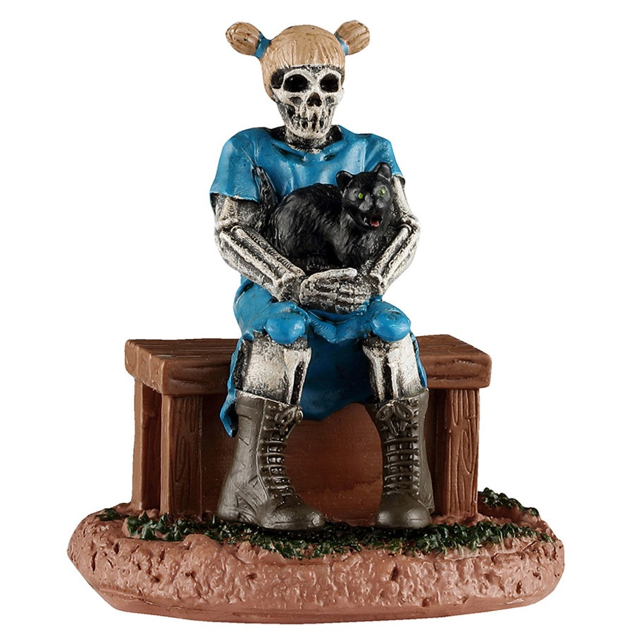 This image shows what this Lemax Spooky Town figurine looks like on a white background. It was designed to look like a skeletal girl sitting on a wooden bench. She has blonde hair in pigtails and she's wearing a tattered blue dress with brown combat boots. In her arms, there's a large fluffy, black cat with green eyes.