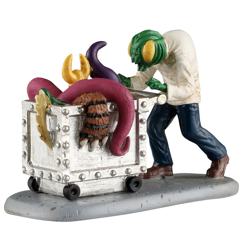 This image shows what this Lemax Spooky Town figurine looks like on a white background. It was designed to look like a green alien with yellow eyes wearing a white doctor's coat, blue jeans and brown shoes. He's pushing a silver cart full of tentacles, claws and other weird monster parts.