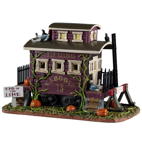 This image shows what this Spooky Town table accent looks like on a white background. It looks like the end of a rundown, very old train. There are crows on the roof and rotting pumpkins on the ground. There's a sign that reads "End of the Line."