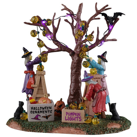 This image shows what this Lemax Spooky Town table accent looks like on a white background. A group of three witches are decorating a bare tree with pumpkin lights and Halloween ornaments.