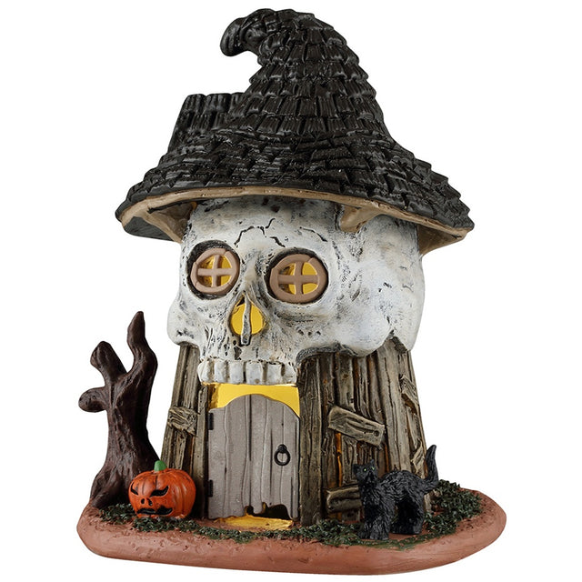 This image shows what this Lemax Spooky Town table accent looks like on a white background. It was designed to look like a wooden house with a skull wearing a witch's hat for a roof. You can see the yellow glow from inside through the two eye windows and the door.