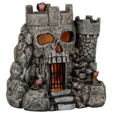This image shows what this Lemax Spooky Town table accent looks like on a white background. It was designed to look like a stone castle. The main part of the castle looks like a skull. There's a black gate where the skull mouth is. There's an orange glow inside. This castle has a vulture on the roof and on both sides of the door.
