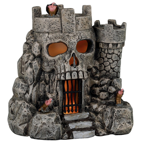 This image shows what this Lemax Spooky Town table accent looks like on a white background. It was designed to look like a stone castle. The main part of the castle looks like a skull. There's a black gate where the skull mouth is. There's an orange glow inside. This castle has a vulture on the roof and on both sides of the door.