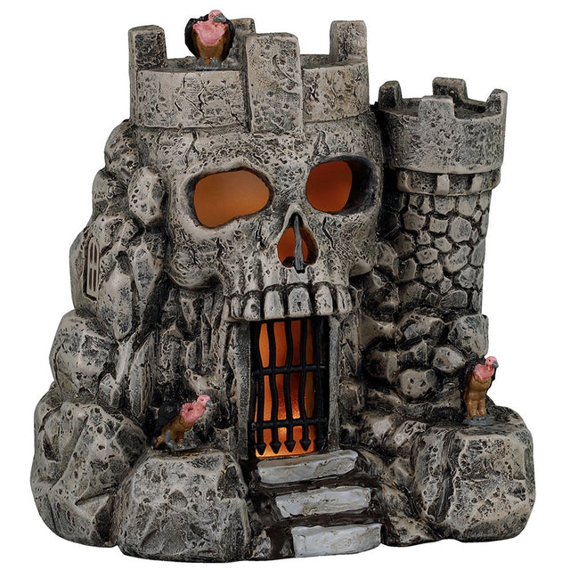 This image shows what this Lemax Spooky Town table accent looks like on a white background. It was designed to look like a stone castle. The main part of the castle looks like a skull. There's a black gate where the skull mouth is. There's an orange glow inside. This castle has a vulture on the roof and on both sides of the door.