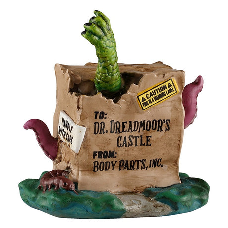 This image shows what this Lemax Spooky Town accessory looks like on a white background. It was designed to look like a dilapidated cardboard box with a green hand punching through the top. There are two maroon colored tentacles coming out the sides and a large, brown centipede coming out the bottom. The box reads "To: Dr. Dreadmoor's Castle. From: Body Parts, Inc. CAUTION: This is warning label."