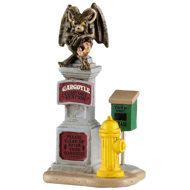 This image shows what this Lemax Spooky Town accessory looks like on a white background. It was designed to look like a patch of sand with a yellow fire hydrant, a tall, stone sign and a green baggy box. The stone reads "Gargoyle Comfort Station. Please clean up after your gargoyle." On top of the sign, there's a brown gargoyle with yellow eyes holding a pink donut.