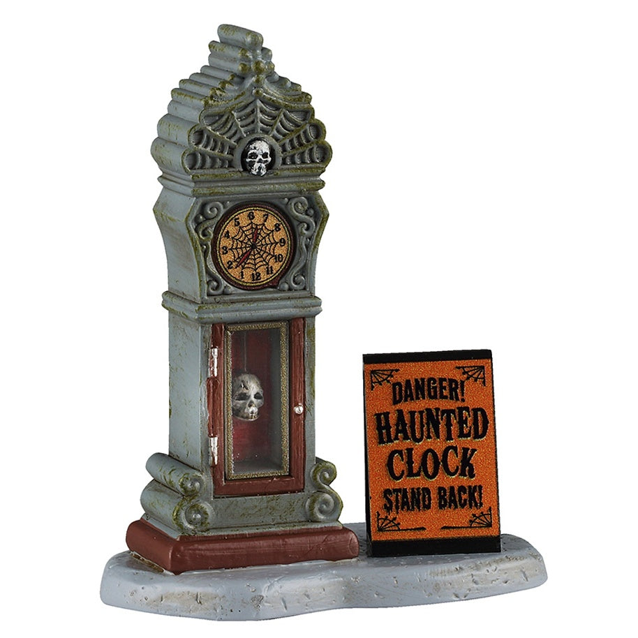 This image shows what this Lemax Spooky Town accessory looks like on a white background. It was designed to look like an old, slightly dirty antique clock. The top looks like spiderwebs with a skull in the center. The face of the clock is also a spiderweb. The base of the clock is glass and you can see a skull inside. There's an orange sign next to it that reads "DANGER! Haunted clock. Stand back!"
