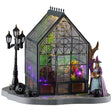 This image shows what this Lemax Spooky Town table accent looks like on a white background. It was designed to look like a greenhouse with plants inside and a witch standing outside.