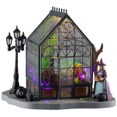 This image shows what this Lemax Spooky Town table accent looks like on a white background. It was designed to look like a greenhouse with plants inside and a witch standing outside.