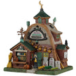 This image shows what this Lemax Spooky Town lighted building looks like on a white background. It was designed to look like a U-shaped building. There's a witch standing at the front door. There's an outdoor stall with lots of herbs and potions on display. There are three tombstone signs on the sidewalk near the front door.