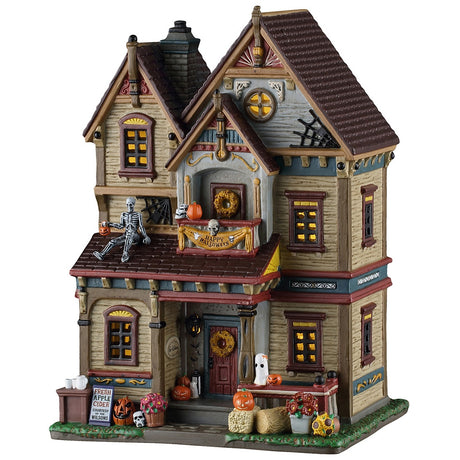 This image shows what this Lemax Spooky Town lighted building looks like on a white background. It was designed to look like a tall, old-timey home. Beautiful autumn arrangements of sunflowers and chrysanthemums perch below the porch while seasonal bales of hay and grinning jack-o’-lanterns line the way to the door. Beneath the awning, a colorful wreath entertains guests while they wait. Around the corner, fresh apple cider is available on the lawn.