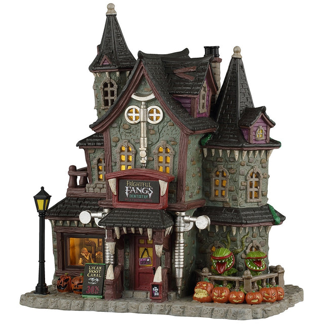 This image shows what this Lemax Spooky Town lighted building looks like on a white background. It was designed to look like a spooky, Victorian-era styled mansion. Above the front door, there's a sign that reads "Frightful Fangs Dentistry." There's also a sign off to the left of the front door that reads "Lycan Root Canal 30% off." There's a window in the front you can look into and see someone in the dentist's chair. Off to the right, there are plants with mouths and lots of jack-o-lanterns.