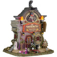 This image shows what this Lemax Spooky Town lighted building looks like on a white background. It was designed to look like a tall building shaped like a headstone. The sign reads "MORTICIAN MAX. DEATH DAY SERVICES" and above it, there's a Victorian-era looking skeleton man tilting his top hat. There's a skeleton with a skeleton standing near the front door.
