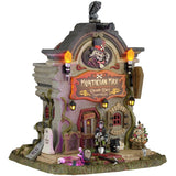 This image shows what this Lemax Spooky Town lighted building looks like on a white background. It was designed to look like a tall building shaped like a headstone. The sign reads "MORTICIAN MAX. DEATH DAY SERVICES" and above it, there's a Victorian-era looking skeleton man tilting his top hat. There's a skeleton with a skeleton standing near the front door.