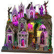 This image shows what this Lemax Spooky Town facade looks like on a white background. It's designed to look like a cemetery on a big hill. There are mausoleums and crypts and tombstones. Parts of it are lit with a purple light. You can see monsters, skeletons and coffins in the doorways. The Grim Reaper stands at the top of the hill holding a scythe.