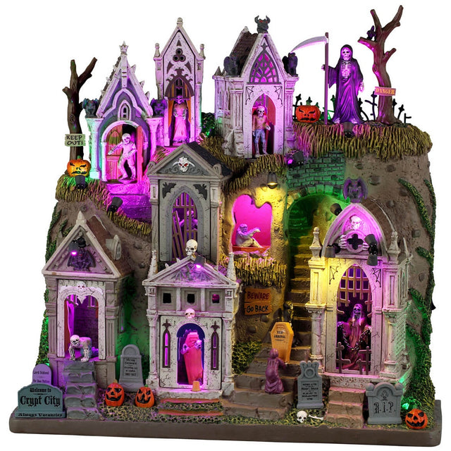 This image shows what this Lemax Spooky Town facade looks like on a white background. It's designed to look like a cemetery on a big hill. There are mausoleums and crypts and tombstones. Parts of it are lit with a purple light. You can see monsters, skeletons and coffins in the doorways. The Grim Reaper stands at the top of the hill holding a scythe.