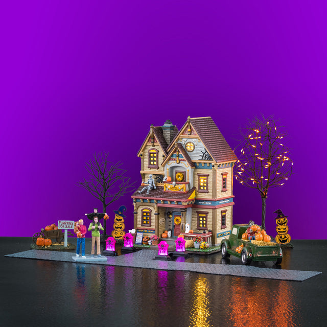 This image shows what this Lemax Spooky Town house set looks like on a purple background. It was designed to look like a house decorated for Halloween. There are purple glowing tombstones, two stacks of jack-o-lanterns, a green pickup truck with a skeleton and a stack of pumpkins in the bed, two bare trees - one has orange lights, a stack of pumpkins for sale and the Wilson family. The mom is holding a baby dressed as a bear. The dad has a kid dressed as a bat sitting on his shoulders.