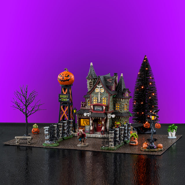 This image shows what this Lemax Spooky Town house set looks like with a purple background. It comes with a pebble road, a black tree with orange lights, a small bare tree, a jack-o-lantern lamppost, a stone bench, a werewolf, three small plants, a Spooky Town water tower, six black gate sections, the Frightful Fangs Dentistry lighted building, a replacement light and an adaptor.