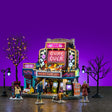 This image shows what this Lemax Spooky Town house set looks like on a purple background. It's designed to look like an arcade made out of games. There's a big screen that reads "GAME OVER." There are zombies crawling and walking around outside. There are two lampposts and two bare trees. One has orange lights.