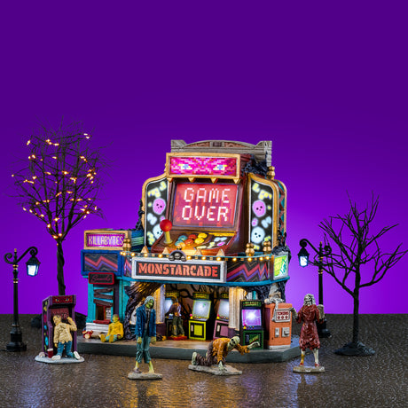 This image shows what this Lemax Spooky Town house set looks like on a purple background. It's designed to look like an arcade made out of games. There's a big screen that reads "GAME OVER." There are zombies crawling and walking around outside. There are two lampposts and two bare trees. One has orange lights.