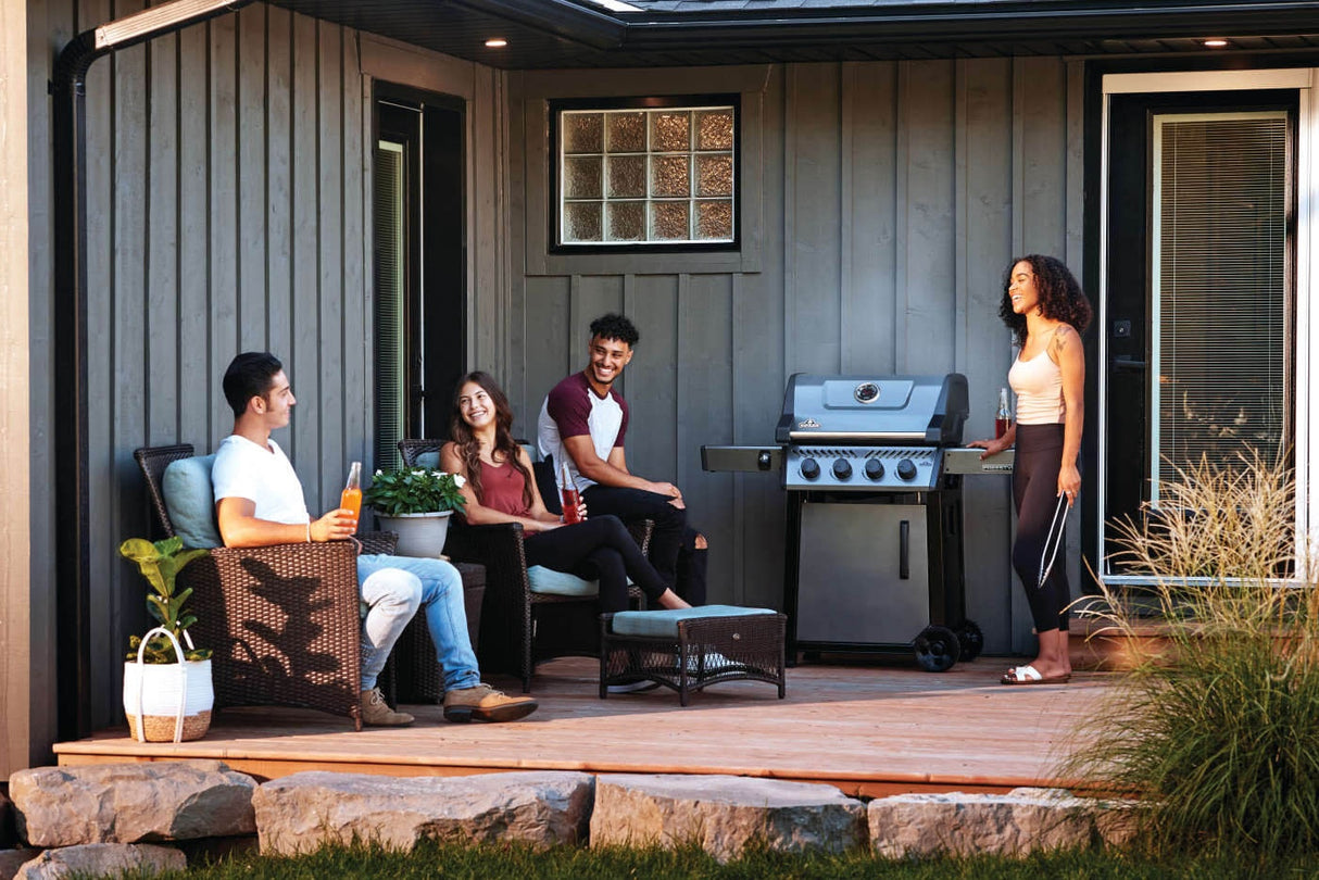 This is a lifestyle image showing a woman standing near the grill holding tongs while she talks to three people who are sitting on the back patio with her.