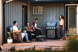 This is a lifestyle image showing a woman standing near the grill holding tongs while she talks to three people who are sitting on the back patio with her.