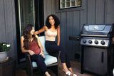 This is a lifestyle image showing the grill along the side of a house, while two women sit and chat.