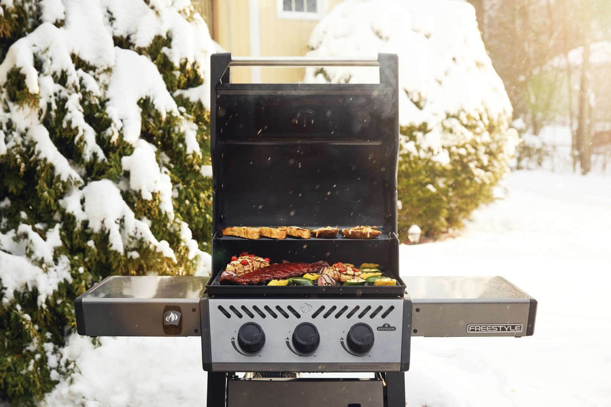 This is a lifestyle image showing this grill in use during winter. There's food grilling on the top rack, as well as on the bottom rack. There's snow on the ground and on the bushes behind the grill.