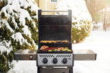 This is a lifestyle image showing this grill in use during winter. There's food grilling on the top rack, as well as on the bottom rack. There's snow on the ground and on the bushes behind the grill.