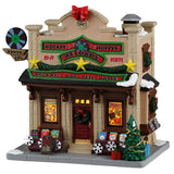 This image shows what this Lemax building looks like on a white background. It was designed to look like a tall record shop. There's a red bow and fake film reel being used as decorations on the roof. The roofline is decorated with garlands, wreaths and gold stars. Near the front door, there are wooden containers of Christmas vinyl and albums. There's a sign above the front door that reads "LPs & 45s, Buy, Sell, Trade."