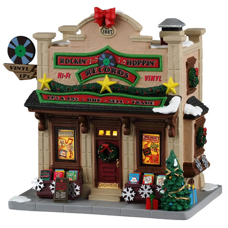 This image shows what this Lemax building looks like on a white background. It was designed to look like a tall record shop. There's a red bow and fake film reel being used as decorations on the roof. The roofline is decorated with garlands, wreaths and gold stars. Near the front door, there are wooden containers of Christmas vinyl and albums. There's a sign above the front door that reads "LPs & 45s, Buy, Sell, Trade."