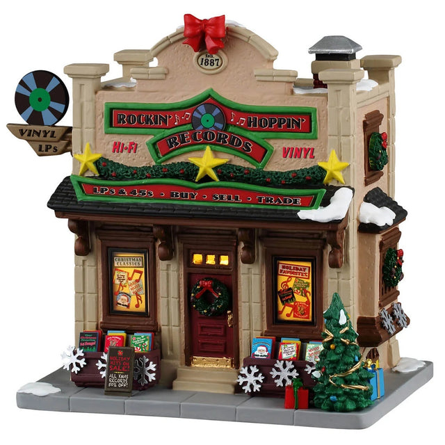 This image shows what this Lemax building looks like on a white background. It was designed to look like a tall record shop. There's a red bow and fake film reel being used as decorations on the roof. The roofline is decorated with garlands, wreaths and gold stars. Near the front door, there are wooden containers of Christmas vinyl and albums. There's a sign above the front door that reads "LPs & 45s, Buy, Sell, Trade."